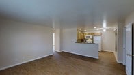 This single-floor space is designed with an open-concept layout that seamlessly connects the living area to the kitchen, creating a cohesive and functional environment. The living area is generously proportioned, featuring warm hardwood-style flooring that extends throughout the space, complemented by neutral-toned walls and white baseboards that lend a clean, understated aesthetic.

The kitchen is positioned at the far end of the open floor plan and is outfitted with medium-toned wood cabinetry, a dark stone-style backsplash, and a raised breakfast bar that provides both a casual dining surface and a natural visual division between the kitchen and living areas. White appliances, including a refrigerator, electric range, and over-the-range microwave, are neatly integrated into the cabinetry. Recessed lighting illuminates the kitchen, while a ceiling fan is visible nearby, enhancing ventilation and comfort.

A textured ceiling adds subtle visual interest throughout the space, and a floor vent indicates a central HVAC system. A doorway at the far left suggests access to additional rooms, hinting at further functional spaces within this well-organized, single-level layout.