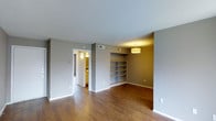 This single-floor space in Irving presents a straightforward and functional layout with a modern, understated aesthetic. The main living area is characterized by rich hardwood flooring that runs throughout, complementing the neutral gray walls and white trim to create a clean, contemporary look. A built-in shelving unit is recessed into one wall, offering practical display and storage space while adding architectural interest to the room. A drum-style pendant light fixture overhead provides warm, focused illumination, enhancing the inviting atmosphere of the space. Ceiling-mounted HVAC vents indicate a central air system throughout. A doorway visible from the main area leads into an adjacent room, where warm lighting suggests a well-lit interior space, potentially a bedroom or walk-in area. White baseboards frame the perimeter of the room, adding a crisp contrast against the darker flooring. The overall design is simple and functional, with enough neutral tones and natural material choices to accommodate a variety of interior styles. The single-floor configuration ensures easy accessibility and a cohesive flow throughout the entire space.