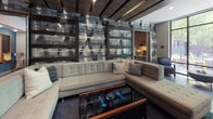 Located at 401 W Harnett St in Raleigh, NC, this single-floor amenity space delivers a sophisticated urban atmosphere through a thoughtfully designed layout with distinct zones for socializing, relaxing, and entertaining. The entrance features black-framed glass doors and polished concrete floors that extend throughout, grounding the space with an industrial-modern aesthetic.

The lounge area showcases bold geometric-patterned wall panels, exposed wood ceiling beams, and tufted sectional seating centered around a sleek glass coffee table. Adjacent to this, a cozy media lounge offers rich brown leather seating, a wall-mounted flat-screen TV framed in a metallic surround, and a sculptural multi-globe chandelier. Wood-paneled accent walls add warmth and texture throughout both areas.

The kitchen and bar zone features a long white quartz island with bar-height seating, pendant lighting with teardrop fixtures, dark cabinetry, and open industrial-pipe shelving against an exposed brick wall — ideal for catering and social gatherings. A dedicated gaming area rounds out the entertainment offerings, complete with a billiard table, high-top bar counter, mounted TVs, and linear LED ceiling accents. A bright café nook with floor-to-ceiling windows frames views of the surrounding streetscape, providing an inviting spot for casual seating. A glassed-in conference or meeting room adds functional versatility to this dynamic community space.