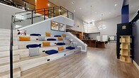 This contemporary community clubhouse is designed to foster social interaction and relaxation through a thoughtfully curated, open-concept layout. The centerpiece of the main level is a striking tiered seating area reminiscent of an amphitheater, finished in light wood tones and adorned with navy, amber, and gray cushions — ideal for gatherings, presentations, or casual conversation. Adjacent to it, a sleek bar-height counter with white quartz tops and warm wood cabinetry anchors the space beneath soaring ceilings accented by angular LED lighting strips and a sculptural starburst chandelier. Longhorn skull mounts and Western-inspired artwork add a regional character throughout. A comfortable lounge area features a tufted sectional, leather accent chairs, and a hexagonal-tile fireplace wall flanked by built-in shelving — creating an inviting retreat for residents. The upper mezzanine level, accessed by a glass-railed staircase, offers additional seating and overlooks the main floor. Bold orange and navy accent walls punctuate the neutral gray palette, while light wood-look flooring flows seamlessly throughout. Large windows and a dramatic cityscape mural contribute to the vibrant, urban ambiance of this well-appointed communal space.