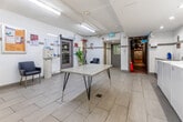 Located at 130 Rosedale Valley Rd in Toronto's Rosedale neighbourhood, this shared laundry facility offers a clean, functional, and well-organized space across a single floor. The layout is divided into two connected areas, providing ample room for residents to complete their laundry needs comfortably.

The main laundry room features a row of front-loading washing machines and top-loading dryers lining the walls, all finished in white and equipped with digital controls for ease of use. Recessed lighting illuminates the space brightly, complemented by fluorescent strip lighting overhead. Large-format light grey floor tiles run throughout, contributing to a clean, modern aesthetic. A central folding table with hairpin legs provides a practical surface for sorting and folding garments.

An adjoining waiting area offers seating with quilted black accent chairs, a bulletin board with posted notices, a small bookshelf, and a payment kiosk for machine operation. Exposed utility piping runs along the ceiling, adding an industrial touch to the otherwise bright and tidy interior. Multiple access doors connect the space to the broader building, including a garage-level entry. Informational signage is clearly posted throughout, ensuring a straightforward and user-friendly experience for all residents.