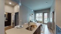 Located at Upper Lakes Estates in Valley Park, Missouri, this single-floor residence offers a thoughtfully designed open-concept layout with contemporary finishes throughout. The kitchen serves as a central focal point, featuring rich dark cabinetry, stainless steel appliances, a mosaic tile backsplash, and a large quartz-topped island with an undermount double sink and pendant lighting — ideal for both meal preparation and casual dining. The kitchen flows seamlessly into the living area, which boasts soaring ceilings, warm wood-look flooring, and generous windows that allow abundant natural light to fill the space. The bedroom is a calm retreat with plush carpeting, a large window with exterior views, and direct access to a well-appointed en-suite bathroom featuring dark wood vanity cabinetry, a glass-enclosed shower, and wood-look tile flooring. A second bathroom includes a full bathtub and shower combination. A spacious walk-in closet with wire shelving provides ample storage. An additional flexible room with large windows and carpet offers versatility as a den or home office. The overall aesthetic blends cool gray tones with warm wood accents, creating a modern yet comfortable ambiance throughout.