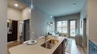 Located at Upper Lakes Estates in Valley Park, Missouri, this single-floor residence offers a thoughtfully designed open-concept layout with contemporary finishes throughout. The kitchen serves as a central focal point, featuring rich dark cabinetry, stainless steel appliances, a mosaic tile backsplash, and a large quartz-topped island with an undermount double sink and pendant lighting — ideal for both meal preparation and casual dining. The kitchen flows seamlessly into the living area, which boasts soaring ceilings, warm wood-look flooring, and generous windows that allow abundant natural light to fill the space. The bedroom is a calm retreat with plush carpeting, a large window with exterior views, and direct access to a well-appointed en-suite bathroom featuring dark wood vanity cabinetry, a glass-enclosed shower, and wood-look tile flooring. A second bathroom includes a full bathtub and shower combination. A spacious walk-in closet with wire shelving provides ample storage. An additional flexible room with large windows and carpet offers versatility as a den or home office. The overall aesthetic blends cool gray tones with warm wood accents, creating a modern yet comfortable ambiance throughout.