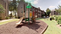 This outdoor amenity space serves as a central gathering hub for the residential community, offering a well-rounded combination of recreational features set against a backdrop of mature trees and multi-story buildings. The playground area is anchored by a multi-level play structure equipped with two slides, climbing panels, and a distinctive green-roofed tower, all set atop a generous rubber mulch safety surface bordered by a curved edging. The layout encourages active play while maintaining a safe and contained environment.

Adjacent to the playground, the swimming pool area is a standout feature of the community. The generously sized pool is complemented by an attached spa, both finished in a clean, light-toned surface that enhances the water's bright appearance. The surrounding concrete deck is outfitted with numerous lounge chairs and shade umbrellas, providing ample space for relaxation.

A covered poolside structure with dark wood-framed columns and stucco walls offers a shaded transition zone between the indoor pool house and the open deck. Large glass doors allow clear sightlines to the pool from within, while wall-mounted lighting and potted plants add a welcoming touch. Pool rules are posted prominently, reflecting an attentive, well-managed amenity space.