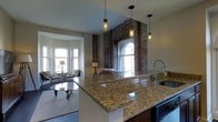 Located at 699 N Broad St in Philadelphia, PA, this single-floor space masterfully combines historic character with contemporary design. The open-concept layout seamlessly connects the kitchen and living areas, creating a cohesive and inviting atmosphere throughout.

The kitchen is a standout feature, anchored by a large granite-topped island with warm gold and cream toning that catches the light beautifully. A sleek, professional-style faucet and modern pendant lighting — featuring industrial cage-style fixtures — hang above the island, adding an on-trend touch to the space. Dark cabinetry provides a rich contrast to the bright countertops.

Exposed brick walls are a defining architectural element, lending the space a raw, historic character that pays homage to the building's heritage. A striking arched window set within the brick framework floods the area with natural light and serves as a dramatic focal point. Additional bay windows in the living area further enhance the brightness of the space, framing views of the surrounding neighborhood.

The living area offers a comfortable, well-proportioned layout with room for seating arrangements centered around a glass coffee table. Neutral tones throughout the walls and flooring keep the aesthetic clean and versatile, allowing the architectural details to take center stage. The overall ambiance is warm, urban, and full of character.
