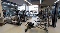 This fitness center offers a comprehensive workout environment housed within a single-floor layout designed to accommodate a variety of exercise routines. The space is characterized by its open floor plan, which provides ample room to move between equipment without feeling cramped.

Natural light floods the interior through large, floor-to-ceiling windows that line the perimeter, creating a bright and energizing atmosphere. The windows also offer views of the surrounding exterior, adding a sense of openness to the workout environment. Overhead, linear fluorescent lighting ensures consistent illumination throughout the space.

The cardio zone is well-stocked with a diverse selection of machines, including treadmills, elliptical trainers, and stationary steppers, arranged to maximize accessibility and traffic flow. A dedicated strength and functional training area features additional equipment such as an abdominal crunch machine and multi-station cable systems, catering to a full-body workout regimen.

Full-length mirrors are mounted along the walls, allowing users to monitor their form during exercise while also visually expanding the space. The clean, light-toned flooring ties the room together and contributes to the facility's modern, professional aesthetic. Overall, this fitness center is thoughtfully designed to support a wide range of fitness goals in a practical and welcoming setting.