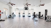 This fitness center offers a spacious, single-floor layout designed to accommodate a full range of exercise activities. The interior is defined by crisp white walls and light hardwood-style flooring that together create a clean, energizing atmosphere throughout the space.

The main workout area is generously proportioned, providing ample room between equipment for comfortable movement and exercise. A row of arched windows along the primary wall floods the space with abundant natural light, enhancing the open and airy feel. Multiple ceiling fans are suspended overhead, ensuring consistent airflow during workouts, while recessed lighting supplements illumination throughout.

The facility is outfitted with a comprehensive selection of equipment, including treadmills, elliptical trainers, stationary and recumbent bikes, a dumbbell rack, and multiple strength-training machines from Life Fitness, covering muscle groups from the upper to lower body. An adjustable weight bench and a punching bag add further versatility to the workout options available.

A large flat-screen TV is mounted from the ceiling, visible from most areas of the room, offering entertainment during exercise sessions. One wall features full-length mirrors that visually expand the space and allow for form monitoring during workouts. A separate entryway area with glass-panel doors connects the fitness floor to an adjacent space, rounding out this well-appointed facility.