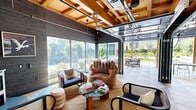 Located at 3705 S Alaska St in Seattle's Columbia City neighborhood, this single-level communal amenity space offers a well-appointed blend of covered and open-air environments designed for gathering and relaxation. The centerpiece of the space is a covered indoor-outdoor lounge area defined by a striking exposed wood beam ceiling with a retractable glass garage door that opens fully to the exterior courtyard, seamlessly connecting interior and outdoor living. Large floor-to-ceiling windows flood the lounge with natural light, while eclectic seating arrangements — including woven accent chairs and a glass-top coffee table — create a welcoming, design-forward atmosphere.

Flowing outward, a partially covered patio features a generous L-shaped sectional sofa with plush cushions, oriented toward lush greenery and framed by the same warm wood ceiling structure strung with ambient bistro lighting. The geometric-patterned large-format tile flooring extends throughout the covered and uncovered patio zones, providing visual continuity. An outdoor kitchen equipped with a stainless steel grill and cabinetry anchors one corner, alongside communal dining tables and seating. The open courtyard beyond showcases a well-maintained turf lawn, sculpted planting beds with trees and perennial plantings, and additional seating areas, all surrounded by the multi-story building facades that enclose the space.