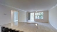 This single-floor residence offers a clean, open layout filled with natural light and thoughtfully appointed finishes throughout. The kitchen features a generous granite countertop with attractive speckled detailing, providing ample workspace and a seamless transition into the open living area beyond. Crown molding runs along the ceiling, adding a refined architectural detail that elevates the overall aesthetic of the space. The living area is carpeted and bathed in natural light streaming through large windows that frame picturesque views of lush, mature trees. A pendant light fixture is centrally positioned overhead, offering both functional illumination and a subtle design accent. A glass-paneled door leads directly to a private balcony, extending the living space outdoors and reinforcing the connection to the surrounding greenery. An adjacent room, visible through an open doorway, offers additional flexible living space and continues the light, neutral color palette found throughout the unit. The walls are finished in soft, warm neutral tones that complement the bright, airy atmosphere. Overall, the space combines practical design with comfortable, inviting details, making it well-suited for everyday living.