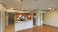 This single-floor space showcases an open-concept layout that seamlessly integrates the kitchen with the surrounding living areas. The kitchen is equipped with stainless steel appliances, including a French door refrigerator, electric range, and built-in microwave, all set against warm medium-toned wood cabinetry. A tile backsplash adds a subtle design element, while granite countertops extend across the workspace and onto a peninsula that provides additional prep space and casual seating potential. Two pendant lights hang above the peninsula, offering focused task lighting with a decorative touch. The flooring throughout is rich hardwood, adding warmth and continuity to the open space. Crown molding runs along the upper walls, lending a refined architectural detail to the interior. A built-in open shelving unit is positioned near a corridor, offering functional display and storage space while visually defining the transition between areas. Ceiling fans are visible overhead, contributing to both comfort and air circulation. The neutral wall tones create a clean, versatile backdrop throughout the space.