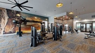 Located in Clermont, FL, this fitness center offers a comprehensive, amenity-rich workout facility spread across a single, generously sized floor. The main workout area is defined by soaring ceilings, stylish gold pendant lighting, and large-blade ceiling fans that enhance airflow and comfort. Patterned carpet tile flooring spans the strength training zone, where an extensive array of cable machines, multi-station weight systems, benches, a functional training rig with TRX suspension straps, and a boxing/heavy bag station are thoughtfully arranged. A striking floor-to-ceiling scenic mural adds a bold visual focal point, while an exposed brick accent wall contributes an industrial warmth to the space. A cardio zone along the perimeter features a row of treadmills and ellipticals positioned in front of large windows, flooding the area with natural light and offering views of lush outdoor surroundings including palm trees and a pool terrace. A separate studio room, accessed through glass-panel doors, provides a dedicated space for cycling bikes and group fitness, with hardwood-style flooring, a large ceiling fan, and a smart mirror for interactive workouts. The entry vestibule also features a smart mirror, rounding out a modern, technology-forward fitness experience.