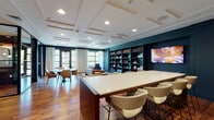This thoughtfully designed single-floor workspace combines refined aesthetics with versatile functionality across several distinct zones. Rich hardwood flooring runs throughout, complementing deep teal accent walls and a cohesive dark-toned color palette that lends the space a polished, professional atmosphere.

The open main area serves as a central hub, anchored by a large communal bar-height table with upholstered seating, ideal for collaborative work or informal gatherings. A dramatic built-in bookcase wall, finished in the same deep teal, provides both visual impact and practical storage, flanked by a wall-mounted display screen. Recessed lighting and a coffered ceiling overhead add architectural detail and warmth.

A glass-partitioned conference room offers a more private setting, featuring a striking live-edge wood table surrounded by upholstered rolling chairs, a wall-mounted display, and built-in shelving with curated décor. The glass walls and black steel framing maintain visual openness while providing acoustic separation.

Additional glass-enclosed rooms accommodate smaller group meetings or focused work, furnished with round tables and accent chairs. Near the entrance, a lounge area with mid-century inspired furniture and patterned rug creates a welcoming, relaxed setting. Large windows throughout allow natural light to filter in, enhancing the space's inviting ambiance.