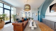 Located at 1720 Nick Nuccio Parkway in Tampa, FL, this thoughtfully designed single-floor living space offers a cohesive and vibrant atmosphere that balances eclectic style with functional comfort. The open layout of the living room is anchored by warm cork-tone flooring and crisp white walls, punctuated by a striking teal accent wall featuring a dramatic arched alcove — a standout architectural element that lends a bold, artistic focal point to the room. A wall-mounted flat-screen TV and a built-in wet bar with dark cabinetry and a beverage refrigerator are seamlessly integrated into this alcove, adding both sophistication and utility. Large black-framed windows line the perimeter, allowing generous natural light to flood the space and offer open views of the surrounding Tampa skyline. An oversized woven pendant light hangs centrally, complementing the warm, layered lighting provided by decorative wall sconces. The seating area is elegantly furnished with a tufted cognac leather Chesterfield sofa, a sculptural white coffee table, and an eclectic mix of accent chairs upholstered in bold botanical and animal prints. Ornate arched mirrors and lush tropical plants add depth and a sense of organic warmth. The overall ambiance is vibrant yet refined, blending contemporary design with artistic character.