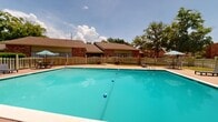 Located at 2335 Atkinson Rd in Biloxi, Mississippi, this single-level outdoor amenity area offers a well-maintained recreational space designed for relaxation and active leisure. At the heart of the space is a generously sized outdoor swimming pool with clear, blue-tinted water, framed by a wide concrete pool deck that provides ample room for sunbathing and socializing. The deck is furnished with lounge chairs, tables, and shade umbrellas, creating a comfortable setting for outdoor enjoyment under the Gulf Coast sun. A white wooden privacy fence encloses a portion of the pool area, lending a sense of seclusion while complementary mature trees provide natural shade along the perimeter. Adjacent to the pool, a full-size tennis court enclosed by chain-link fencing adds an additional layer of recreational opportunity to the property. The surrounding brick-and-siding building architecture, characteristic of the region, frames the space and contributes to its welcoming neighborhood feel. Lush greenery and well-kept landscaping enhance the overall ambiance, making this outdoor area an inviting retreat for residents and guests alike.