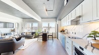 This thoughtfully designed urban residence offers a seamless open-concept layout that integrates the living, dining, and kitchen areas into one cohesive, light-filled space. Wide windows line the main living area, flooding the unit with natural light and framing expansive city views. The kitchen is a standout feature, boasting crisp white shaker cabinetry, a striking marble-veined backsplash, light quartz countertops, stainless steel appliances including a dishwasher and built-in oven, and a deep undermount sink — all finished with sleek black hardware. Warm wood-tone flooring flows throughout, complementing the exposed concrete ceiling elements that lend an industrial-chic character to the space. The living area offers generous room for seating, anchored by large windows overlooking the urban skyline. The bedroom is bright and serene, featuring a city-facing window, high ceilings with a concrete finish, and track lighting. The en-suite bathroom is elegantly appointed with a dual-sink vanity, white cabinetry, a walk-in glass shower with marble-style tile surround, and dark accent fixtures. A dedicated in-unit laundry nook with overhead cabinetry adds everyday convenience. The overall aesthetic blends contemporary design with industrial accents for a refined urban living experience.
