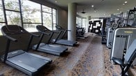 This fitness center is designed to provide a comprehensive workout environment within a single, open-plan floor. The space is lined with large windows along one side, allowing abundant natural light to fill the room while offering views of the surrounding greenery — an inviting backdrop for cardio sessions. A row of treadmills is prominently positioned to take advantage of these views, supplemented by elliptical trainers and additional cardio equipment arranged throughout the space. Structural columns punctuate the layout, adding an architectural element without disrupting the flow of the floor plan. A ceiling fan overhead contributes to air circulation, maintaining comfort during workouts. The flooring features a richly patterned, decorative carpet that adds a layer of visual warmth and sophistication uncommon in typical fitness spaces. Toward the rear of the room, a bold geometric shelving unit with a black-and-white pattern serves as a striking design focal point. Recessed lighting in the ceiling complements the natural light, ensuring the space remains well-lit throughout the day. Overall, the fitness center balances function and aesthetics, offering a well-appointed environment suited for a variety of cardiovascular training routines.