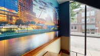 This distinctive entryway and stairwell space makes a strong first impression through a carefully considered combination of color, art, and architecture. The interior is defined by striking blue accent walls paired with warm cream tones and rich wood trim details that run along the staircase landing, creating a layered and visually engaging palette. A standout feature is the large-scale photographic cityscape mural — depicting a dramatic urban skyline with a striking cable-stayed bridge — that wraps across one wall, adding depth and a bold artistic focal point to the space. The staircase itself is constructed with steel railings and carpeted treads in a muted multi-tone pattern, lending both an industrial and functional character. Floor-to-ceiling glass panels along the street-facing side invite abundant natural light and offer open views to the surrounding urban streetscape, connecting the interior to the neighborhood beyond. The entry level features tile flooring and multiple unit access doors finished in coordinating blue-gray tones, maintaining design continuity throughout. An elevator is also present, providing convenient vertical access alongside the staircase. The overall ambiance is urban and contemporary, balancing practical circulation design with thoughtful decorative elements that give the space a distinct and welcoming character.