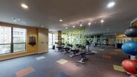 This fitness center is a comprehensive, single-floor facility designed to support a full range of workout routines. The space is accessed through a glass-paneled entry vestibule, which opens into a generously sized main floor with colorful multi-toned carpet tiles that add a vibrant, energetic aesthetic throughout.

The cardio zone features multiple treadmills and elliptical machines positioned near large floor-to-ceiling windows, allowing natural light to fill the space and offering views of the surrounding exterior. A dedicated spinning area houses a row of stationary cycling bikes, set against a decorative green patterned partition and mirrored wall that amplifies the sense of space.

The strength training section is well-appointed with a variety of selectorized machines for targeted muscle groups, alongside a free weights station equipped with a full rack of dumbbells. Wall-mounted resistance bands and suspension training straps provide additional options for functional fitness. Motivational wall art and decals contribute to an encouraging atmosphere throughout.

Track lighting and recessed ceiling fixtures ensure consistent, well-distributed illumination across all workout zones. The open layout promotes easy movement between equipment areas, and the mirrored walls further enhance the sense of openness. Overall, this fitness center delivers a clean, modern environment with the amenities needed for a complete and effective workout experience.