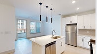 Located at 19 Harnett St in Wilmington, NC, this modern 2-bedroom, 1-bathroom residence offers a thoughtfully designed single-floor layout with clean finishes throughout. The open-concept kitchen and living area serves as the heart of the home, anchored by a central island with quartz countertops, a deep stainless steel single-basin sink, and a matte black faucet. Sleek white cabinetry lines the walls alongside stainless steel appliances, including a refrigerator, microwave, and dishwasher. A tile backsplash and trio of cylindrical pendant lights add a contemporary touch, while light wood-style flooring and access to a private balcony bring warmth and natural light to the space. A carpeted hallway connects the living area to the bedrooms, each finished with soft carpet, ceiling fans, and windows that invite natural light. The primary bedroom includes direct access to a dedicated walk-in closet fitted with wire shelving systems. The bathroom is finished in a crisp, all-white palette, featuring a glass-enclosed walk-in shower, white vanity with quartz countertop, and light wood-style flooring with matte black hardware accents throughout.