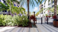 This outdoor amenity space exudes a resort-like atmosphere, thoughtfully designed to combine tropical landscaping with refined architectural details. The grounds are finished with large-format paver walkways in complementary tones, creating a cohesive and polished aesthetic throughout the courtyard.

Towering palm trees and dense tropical plantings — including vibrant red cordyline, broadleaf shrubs, and lush green foliage — frame the space, offering both privacy and a lush, garden-like ambiance. A gated pool area is visible beyond the landscaping, with white cabana structures adding to the resort character of the setting.

A covered outdoor lounge pavilion serves as a central gathering point, featuring a warm wood-paneled ceiling with ceiling fans, stacked stone columns with rich, multi-toned textures, and floor-to-ceiling glass doors that seamlessly connect the interior and exterior. The lounge is furnished with contemporary outdoor seating, including woven accent chairs, cushioned sofas with bold orange and blue pillows, and natural wood stump side tables, creating a welcoming and stylish retreat.

The surrounding multi-story buildings with balconies overlook the courtyard, reinforcing the sense of an enclosed, private oasis. Overall, the space strikes a balance between tropical vibrancy and modern design elegance.
