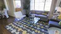This single-floor space showcases a thoughtfully designed interior with a distinctly modern aesthetic. The living area serves as the focal point, bathed in natural light streaming through large windows that frame views of lush greenery outside. The space features polished concrete flooring anchored by a striking geometric area rug in charcoal, yellow, and grey tones that adds a bold, contemporary flair underfoot. Seating is composed of a sleek grey sofa accented with patterned and striped throw pillows, paired with a classic white tufted Barcelona-style chair on a chrome frame — a timeless design piece that elevates the room's sophistication. A transparent glass coffee table with a sculptural base sits at the center, styled with candle arrangements and a small decorative plant, keeping the visual lines open and uncluttered. A tall indoor palm adds a natural, organic element to the corner, softening the modern geometry of the space. The overall ambiance is polished yet inviting, blending clean lines and curated décor to create a space that feels both stylish and functional. The abundant natural light and carefully selected furnishings together contribute to an airy, welcoming atmosphere throughout.