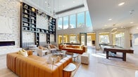 This expansive amenity facility offers a sophisticated blend of modern interior design and resort-style outdoor living across a single sprawling floor. The centerpiece of the interior is a grand lounge area distinguished by soaring double-height ceilings, floor-to-ceiling windows, and an abundance of natural light that floods the space. Pendant globe lighting cascades dramatically overhead, while rich cognac leather sofas and neutral accent chairs are arranged around sleek coffee tables, creating an inviting and stylish atmosphere. A striking floor-to-ceiling built-in shelving unit with textured tile accents anchors one wall, adding architectural depth and visual interest. A linear fireplace set against an artfully tiled feature wall provides a refined focal point. Adjacent areas include a private meeting or dining room with a gold-accented chandelier and glass-panel partitions, and a dedicated game room outfitted with a billiard table, a bold multi-arm brass chandelier, and a large-screen entertainment center. Outdoors, covered patios with ceiling fans offer comfortable seating areas around a built-in fireplace, overlooking a resort-style pool. Lush landscaping with palm trees, manicured greenery, and a spacious paver courtyard with a pergola structure complete the outdoor experience, making this an all-encompassing amenity destination.