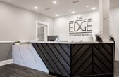 Located at 23 US-1 S in New Brunswick, NJ, the leasing office for The Edge at Raritan Heights is a thoughtfully designed single-floor space that combines contemporary aesthetics with functional professionalism. Upon entry, visitors are welcomed by wide, wood-look tile flooring that extends throughout the space, complemented by two-tone walls in soft gray and white with crisp white wainscoting that lend a polished, refined character.

The reception area serves as the focal point of the space, anchored by a striking front desk featuring a marble-look surface with bold diagonal striping in teal and gray. The branded wall behind the desk prominently displays "The Edge at Raritan Heights" logo, reinforcing a strong sense of identity. Woven globe pendant lights hang at varying heights throughout, adding warmth and a design-forward touch to the overall ambiance.

Recessed tray ceilings with integrated lighting elevate the sense of height and sophistication throughout the open common areas. Glass-enclosed rooms provide transparent views into a conference or meeting space outfitted with a large table and ergonomic seating, ideal for professional consultations.

Hallways lead to additional private rooms and display areas, where property renderings and marketing materials are presented on easels. Decorative geometric mirrors and thoughtful accent pieces complete the interior, creating a welcoming yet informative environment for prospective residents.