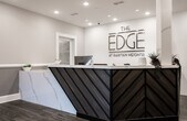Located at 23 US-1 S in New Brunswick, NJ, the leasing office for The Edge at Raritan Heights is a thoughtfully designed single-floor space that combines contemporary aesthetics with functional professionalism. Upon entry, visitors are welcomed by wide, wood-look tile flooring that extends throughout the space, complemented by two-tone walls in soft gray and white with crisp white wainscoting that lend a polished, refined character.

The reception area serves as the focal point of the space, anchored by a striking front desk featuring a marble-look surface with bold diagonal striping in teal and gray. The branded wall behind the desk prominently displays "The Edge at Raritan Heights" logo, reinforcing a strong sense of identity. Woven globe pendant lights hang at varying heights throughout, adding warmth and a design-forward touch to the overall ambiance.

Recessed tray ceilings with integrated lighting elevate the sense of height and sophistication throughout the open common areas. Glass-enclosed rooms provide transparent views into a conference or meeting space outfitted with a large table and ergonomic seating, ideal for professional consultations.

Hallways lead to additional private rooms and display areas, where property renderings and marketing materials are presented on easels. Decorative geometric mirrors and thoughtful accent pieces complete the interior, creating a welcoming yet informative environment for prospective residents.