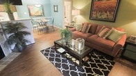 This single-floor residence offers a thoughtfully arranged open-concept layout that connects the living and dining areas, creating a cohesive and functional living environment. The living room is anchored by rich hardwood flooring and features a plush, earth-toned sofa dressed with colorful patterned throw pillows, complemented by a sleek glass-top coffee table resting atop a bold black-and-white geometric area rug. Decorative candle holders and a small fern centerpiece add character to the space, while large framed artwork on the walls contributes warmth and visual interest.

Adjacent to the living room, the dining area accommodates a round table with elegant wrought-iron style chairs beneath a classic pendant light fixture. The soft blue-green wall tones in this zone contrast pleasantly with the warmer hues of the living space, creating distinct yet harmonious areas. A corridor visible in the background suggests additional rooms beyond, hinting at further functional spaces within the home. Overall, the interior presents a well-decorated, inviting atmosphere with a balance of casual comfort and tasteful styling.