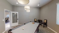 Located at 11427 Porter Ranch Drive in Los Angeles, this single-floor billiard room is thoughtfully designed for leisure and social engagement. The centerpiece of the space is a full-size pool table with a light felt surface and dark wood trim, positioned prominently beneath a trio of elegant drum pendant lights that provide focused, warm illumination. The room is finished with a neutral gray color palette on the walls, complemented by a richly patterned carpet that adds texture and visual interest underfoot. Seating is available throughout, including dark-framed chairs with woven backs and a small bistro-style table, offering comfortable spots for spectators and conversation. Framed artwork adorns one wall, adding a refined touch to the recreational setting. A cue rack mounted on the wall keeps equipment organized and accessible. Lush indoor plants are placed throughout the room, contributing a fresh, lively element to the otherwise neutral interior. Natural light filters in through large windows fitted with horizontal blinds, brightening the space without glare. A glass-paneled door connects the billiard room to an adjacent dining or common area, reinforcing the social, community-oriented character of this well-appointed recreational space.