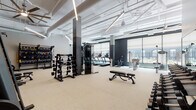 This well-appointed fitness center offers a expansive, open layout designed to accommodate a full range of workout routines. The space features soaring ceilings with exposed ductwork and structural beams, lending an industrial-modern aesthetic that is complemented by sleek linear pendant lighting and large ceiling fans for air circulation. The flooring throughout is a durable, light-toned carpet-style material suited for high-traffic gym use.

The cardio zone is positioned along a wall of large, floor-to-ceiling windows dressed with roller shades, providing ample natural light and views of an outdoor terrace and surrounding landscape. This area houses a row of treadmills, elliptical machines, and stationary bikes. The strength training section is generously equipped with multiple dumbbell racks, a cable machine, a full power rack with barbell and weight plates, adjustable benches, and additional functional training attachments.

A dedicated free weights and heavy lifting area features a striking geometric mural in earthy tones of rust, forest green, and charcoal, accented by a silhouetted athletic figure, adding a motivational and artistic focal point to the space. Stability balls and suspension training equipment round out the functional fitness offerings. Mirror panels are incorporated to enhance spatial perception and support form-checking during workouts. Accent artwork and thoughtful décor details contribute to a polished, contemporary atmosphere throughout.