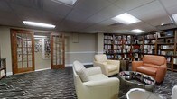 This single-floor library and reading lounge offers a thoughtfully designed space that blends functionality with comfort. The room is defined by expansive built-in bookshelves lining the walls, stocked with a wide variety of books and display items, creating an intellectual and welcoming atmosphere throughout. A patterned carpet with bold, sweeping motifs anchors the seating area, which features plush cream-colored leather armchairs arranged to encourage relaxed reading or quiet conversation. A glass-topped side table adds a touch of contemporary elegance to the lounge area. An additional orange accent chair provides a pop of color and extra seating toward one corner of the room. The space is illuminated by recessed panel lighting set into a suspended ceiling, casting an even, comfortable glow across the room. A classic wood-framed glass-paned door leads to an adjacent room, also lined with bookshelves, extending the library collection further. White chair rail molding runs along the walls, adding a refined architectural detail that complements the overall interior. The layout is open and spacious, making it easy to navigate between the reading areas and shelving. Overall, this library lounge provides a serene and well-curated environment suited for leisurely reading or focused study.