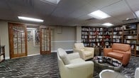 This single-floor library and reading lounge offers a thoughtfully designed space that blends functionality with comfort. The room is defined by expansive built-in bookshelves lining the walls, stocked with a wide variety of books and display items, creating an intellectual and welcoming atmosphere throughout. A patterned carpet with bold, sweeping motifs anchors the seating area, which features plush cream-colored leather armchairs arranged to encourage relaxed reading or quiet conversation. A glass-topped side table adds a touch of contemporary elegance to the lounge area. An additional orange accent chair provides a pop of color and extra seating toward one corner of the room. The space is illuminated by recessed panel lighting set into a suspended ceiling, casting an even, comfortable glow across the room. A classic wood-framed glass-paned door leads to an adjacent room, also lined with bookshelves, extending the library collection further. White chair rail molding runs along the walls, adding a refined architectural detail that complements the overall interior. The layout is open and spacious, making it easy to navigate between the reading areas and shelving. Overall, this library lounge provides a serene and well-curated environment suited for leisurely reading or focused study.