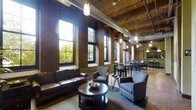 This single-floor space is a standout example of industrial-chic design, seamlessly blending raw architectural elements with refined furnishings. Exposed red brick walls rise to dramatic heights, complemented by warm wooden ceiling planks and visible ductwork that reinforce the loft's industrial character. A series of tall, multi-pane windows line one wall, flooding the interior with abundant natural light and offering views of the surrounding greenery. The open-plan layout is thoughtfully divided into two distinct zones. The lounge area features a rich leather sofa, upholstered armchairs, and a dark wood coffee table, creating an inviting and comfortable gathering space. Adjacent to the lounge, a dining area accommodates multiple tables and chairs, making it well-suited for group meals or meetings. Pendant lighting fixtures hang at varying heights throughout, casting a warm, ambient glow that balances the natural light. Track lighting along the ceiling provides additional illumination. The overall ambiance is one of sophisticated urban warmth — a space where the character of a historic building is preserved and thoughtfully enhanced with modern, functional design.