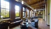 This single-floor space is a standout example of industrial-chic design, seamlessly blending raw architectural elements with refined furnishings. Exposed red brick walls rise to dramatic heights, complemented by warm wooden ceiling planks and visible ductwork that reinforce the loft's industrial character. A series of tall, multi-pane windows line one wall, flooding the interior with abundant natural light and offering views of the surrounding greenery. The open-plan layout is thoughtfully divided into two distinct zones. The lounge area features a rich leather sofa, upholstered armchairs, and a dark wood coffee table, creating an inviting and comfortable gathering space. Adjacent to the lounge, a dining area accommodates multiple tables and chairs, making it well-suited for group meals or meetings. Pendant lighting fixtures hang at varying heights throughout, casting a warm, ambient glow that balances the natural light. Track lighting along the ceiling provides additional illumination. The overall ambiance is one of sophisticated urban warmth — a space where the character of a historic building is preserved and thoughtfully enhanced with modern, functional design.