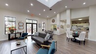Located on Providence Road in Brandon, Florida, this single-story community clubhouse delivers a well-appointed gathering space designed for both comfort and functionality. The interior is defined by its dramatically high ceilings, which lend an open, airy quality to the main lounge area. A striking floor-to-ceiling stacked stone fireplace serves as the room's focal point, anchoring the space with a bold architectural statement alongside a wall-mounted television.

The main lounge is furnished with a mix of contemporary seating, including a tufted gray sofa, mid-century accent chairs in teal and natural wood tones, and sleek metal-framed coffee tables, all set atop durable wood-look plank flooring that runs throughout the building. A skylight overhead supplements the generous natural light streaming in through numerous black-framed glass doors and windows, which offer views of the surrounding tree-lined exterior.

A dedicated meeting or dining nook provides a more intimate setting with a wooden table and upholstered chairs. Adjacent to the main area, a functional kitchen features white cabinetry, extended countertops, and garden-facing windows. A private staff office is also present. The overall ambiance is modern yet welcoming, suitable for community gatherings and everyday use.