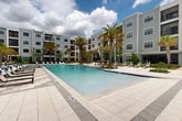 Located at 658 Northwest 120th Terrace in Gainesville, Florida, this outdoor amenity space is designed to deliver a resort-caliber experience within a modern residential community. At the heart of the space is a generously sized swimming pool with crystal-clear blue water, framed by a broad, paver-clad sun deck that provides ample room for lounging and socializing. The pool area is appointed with a curated mix of sleek white contoured chaise lounges and traditional sling-style recliners, many accented with blue cushions, as well as shaded outdoor dining areas complete with umbrellas and contemporary furniture. Towering palm trees are thoughtfully distributed throughout the space, lending a tropical ambiance and natural shade. Lush, layered landscaping with ornamental grasses, shrubs, and tropical plantings softens the hardscape and adds vibrant greenery throughout. A covered pergola structure at the rear of the pool deck offers a shaded retreat and gathering spot. The surrounding multi-story residential buildings feature neutral grey horizontal siding, private balconies, and large windows that overlook the courtyard. Wide, tiled pathways connect the pool area to building entrances, ensuring seamless flow across the entire outdoor space. The overall atmosphere is contemporary, polished, and inviting.