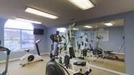 Located at 200 Foxridge Ct in Durham, North Carolina, this single-floor fitness center offers a well-equipped space designed to support a well-rounded workout routine. The room features light blue walls and a neutral carpeted floor, creating a clean and inviting atmosphere. Overhead fluorescent lighting ensures the space is well-lit throughout, complemented by natural light streaming in through a distinctive arched window along one wall, which adds architectural character to the room. A wall-mounted flat-screen television is positioned near the window for entertainment during workouts. The facility is outfitted with a range of cardio equipment, including a treadmill, an elliptical trainer, and a stationary bike, providing diverse options for cardiovascular exercise. A multi-station cable weight machine anchors one side of the room, catering to strength training needs. Floor-to-ceiling mirrors line one wall, visually expanding the space and allowing users to monitor their form. The overall ambiance is functional and practical, making efficient use of the available footprint while delivering a comprehensive set of amenities for residents seeking convenient access to fitness facilities.