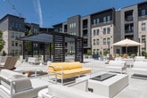 Courtyard 2 is a thoughtfully designed outdoor communal space located in Meridian, Idaho, enclosed by contemporary multi-story buildings featuring a modern architectural palette of brick, horizontal siding, and dark accent panels. The courtyard offers a generous open-air environment bathed in abundant natural sunlight, anchored by a striking wood-and-lattice pergola at its center, strung with overhead bistro lights that lend a welcoming ambiance both day and evening.

The space is organized into distinct activity zones. A wide concrete plaza serves as the main gathering area, furnished with an assortment of outdoor seating arrangements including white lounge sofas with yellow cushions, Adirondack chairs, and high-top dining tables with chairs and umbrellas. A bocce ball court adds a recreational element to the space. Raised concrete planters with young trees and ornamental landscaping provide natural accents throughout.

Surrounding the hardscaped areas, manicured lawn sections offer open green space, complemented by well-maintained landscaping with shrubs and perennials. A paved walkway flanked by greenery connects the courtyard to adjacent areas of the property, ensuring easy pedestrian flow. The enclosing buildings feature private balconies overlooking the courtyard, creating an integrated indoor-outdoor community experience.