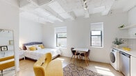 Located at 1045 W Lawrence Ave in Chicago's vibrant Uptown neighborhood, this loft-style studio showcases an open, airy layout with distinctive industrial-inspired architecture. The space is defined by its striking exposed concrete beam ceilings, which lend an authentic loft character while contributing to an impressive sense of vertical height throughout.

The main living area is flooded with natural light through multiple windows offering neighborhood views, creating a warm and inviting atmosphere. The open floor plan seamlessly integrates the sleeping, lounging, and dining areas, making efficient and flexible use of the space. A dedicated closet provides practical storage, and a full-length mirror enhances the sense of openness.

The kitchen is sleekly designed with white cabinetry, a subway tile backsplash, and stainless steel appliances including a range, dishwasher, and refrigerator. A notable feature is the inclusion of an in-unit washer, adding everyday convenience. A window in the kitchen allows natural light to brighten the workspace.

Throughout the unit, polished concrete-style flooring ties the industrial aesthetic together, complementing the crisp white walls. The overall ambiance is modern yet warm, blending urban loft character with clean, contemporary finishes — well-suited for those seeking a stylish and functional home in one of Chicago's most dynamic neighborhoods.