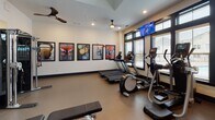 This fitness center offers a comprehensive workout environment designed to support a wide range of exercise routines. The space is thoughtfully laid out across a single floor, with distinct zones dedicated to cardio, strength training, and functional fitness. Large windows fitted with blinds flood the facility with natural light, while recessed lighting and ceiling fans maintain a comfortable atmosphere throughout.

The cardio area is equipped with treadmills, elliptical machines, and a stationary bike, all positioned to face a wall-mounted flat-screen television. A second TV is mounted above the windows, ensuring visibility from multiple areas of the room. Motivational fitness-themed artwork lines the walls, adding energy to the space.

The strength training section features a multi-functional cable machine and a variety of selectorized weight machines suited for both upper and lower body workouts. A full rack of dumbbells spans a mirrored wall, accompanied by storage for stability balls, resistance tools, and kettlebells — providing everything needed for functional and free-weight training.

A glass-paneled partition wall with black steel framing separates a secondary area, which includes additional equipment and access to a utility or restroom space finished with geometric tile. The durable rubberized flooring throughout provides safety and comfort underfoot, making this fitness center a well-rounded facility for users of all fitness levels.