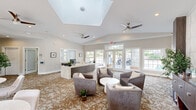 Located at 300 Palmetto Park Blvd in Lexington, South Carolina, this single-floor community clubhouse offers a well-appointed and thoughtfully designed gathering space. The centerpiece of the facility is a grand lobby and lounge area distinguished by soaring vaulted ceilings with a prominent skylight that floods the space with abundant natural light. Dual ceiling fans maintain comfortable airflow throughout. The lounge is furnished with curved, contemporary swivel chairs arranged around a round white coffee table, set atop a richly patterned carpet that anchors the room with warmth and texture. A sleek reception desk with stone accents anchors one side of the lobby, providing a professional welcome point for visitors. Adjacent to the lounge, a stylish dining area features round marble-topped tables with upholstered chairs, positioned beside large French doors that open to views of an outdoor pool, allowing natural light to pour in. A separate office area with vaulted ceilings, large windows, and a built-in stone-topped desk provides a functional workspace. Decorative wall art, lush greenery, a wood-paneled accent wall, and a beverage station add to the refined, resort-inspired ambiance that defines this inviting community hub.