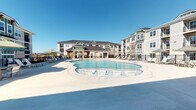This outdoor amenity area centers around a generously sized freeform swimming pool with a shallow wading section equipped with built-in seating, making it suitable for a range of uses. The pool deck is finished in smooth concrete and offers ample space for sunbathing, with multiple lounge chairs and umbrella-shaded seating areas distributed throughout.

A covered pavilion anchors one side of the pool area, constructed with warm wood timber framing and distinctive stacked stone columns that add a refined, lodge-inspired aesthetic. Ceiling fans are mounted overhead within the pavilion, providing ventilation during warm weather. The covered space includes cushioned wicker lounge chairs and side tables, creating a shaded retreat adjacent to the water.

The surrounding multi-story residential buildings feature a combination of fiber cement siding in green and gray tones, stacked stone accents, and private balconies that overlook the pool courtyard. Black metal fencing defines the perimeter of the pool area, with gated access points for security. The open layout of the deck ensures unobstructed views across the water toward the central clubhouse structure, which features a prominent gabled roofline and timber entryway. Under clear skies, the entire space exudes a bright, resort-inspired atmosphere.
