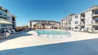 This outdoor amenity area centers around a generously sized freeform swimming pool with a shallow wading section equipped with built-in seating, making it suitable for a range of uses. The pool deck is finished in smooth concrete and offers ample space for sunbathing, with multiple lounge chairs and umbrella-shaded seating areas distributed throughout.

A covered pavilion anchors one side of the pool area, constructed with warm wood timber framing and distinctive stacked stone columns that add a refined, lodge-inspired aesthetic. Ceiling fans are mounted overhead within the pavilion, providing ventilation during warm weather. The covered space includes cushioned wicker lounge chairs and side tables, creating a shaded retreat adjacent to the water.

The surrounding multi-story residential buildings feature a combination of fiber cement siding in green and gray tones, stacked stone accents, and private balconies that overlook the pool courtyard. Black metal fencing defines the perimeter of the pool area, with gated access points for security. The open layout of the deck ensures unobstructed views across the water toward the central clubhouse structure, which features a prominent gabled roofline and timber entryway. Under clear skies, the entire space exudes a bright, resort-inspired atmosphere.