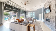 Located in Parker, Colorado, this single-story community clubhouse offers a thoughtfully designed gathering space that seamlessly blends indoor comfort with outdoor recreation. The main living area is anchored by a dramatic floor-to-ceiling stone fireplace feature wall, complete with a built-in linear fireplace and a mounted flat-screen TV. Coordinating wicker and upholstered seating arrangements, patterned area rugs, and open metal shelving with curated decor and board games create an inviting, social atmosphere throughout the space. Soaring ceilings with dual ceiling fans promote airflow and enhance the sense of openness, while polished concrete flooring ties the modern-rustic aesthetic together. Large glass garage-style doors open fully to the outdoor pool deck, allowing natural light to flood the interior and providing effortless indoor-outdoor flow. The game room features a full-size ping-pong table, large windows overlooking the pool, and a small prep counter, making it ideal for active recreation. Outside, an expansive pool deck lined with lounge chairs and shaded cabanas surrounds a resort-style pool with water fountains, set against the backdrop of the attractively landscaped residential community. Stone exterior accents and well-maintained green spaces complete the polished, amenity-rich environment.