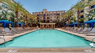 Located at 300 N Beaudry Ave in downtown Los Angeles, this expansive outdoor amenity space is designed to deliver a resort-style experience within a vibrant urban setting. The centerpiece is a large, gleaming swimming pool with crystal-clear blue water, framed by wide stone-tiled pool decks ideal for lounging and socializing. Rows of cushioned chaise lounges are arranged along the poolside, offering ample space to relax under the Southern California sun. Bright blue canopy cabanas are positioned throughout the courtyard, providing shaded retreats for residents and guests. Tall, swaying palm trees are planted throughout the space, lending a tropical ambiance and natural privacy. Raised planter beds burst with vibrant red flowers and lush greenery, adding color and visual texture to the hardscaped surroundings. A pergola structure at one end of the pool provides additional shaded seating, while decorative hand-painted ceramic pots accent entryways and garden corners. Wrought-iron gated entrances with elegant pillar detailing offer controlled access to the courtyard. The multi-story residential buildings surrounding the space feature Mediterranean-inspired architecture with arched windows, warm stucco facades, and private balconies overlooking the courtyard below, completing this well-appointed outdoor retreat.