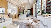 Located at 1140 Harrison Street in San Francisco's SoMa neighborhood, this elegantly designed lobby and amenity space offers a refined first impression through its contemporary architectural design. The interior is anchored by a striking marble reception desk with a veined stone finish, complemented by large-format ceramic tile flooring and soaring ceilings that lend an open, airy feel throughout. Warm wood cabinetry and brass-accented pendant lighting add a layer of sophistication, while curated sculptural artwork and framed pieces contribute to the gallery-like ambiance. A dedicated consultation area features marble-topped workstations paired with display screens, providing a functional yet polished environment. Floor-to-ceiling windows and glass doors flood the interior with abundant natural light and offer seamless visual connectivity to the outdoors. The space transitions to a spacious outdoor courtyard, where large-format stone pavers, raised planter beds with lush greenery, a decorative water fountain feature, and tall palm trees create a tranquil urban retreat. Seating areas with modern furnishings are thoughtfully arranged throughout the courtyard, offering multiple settings for relaxation or casual gatherings. Together, the interior and exterior areas form a cohesive, upscale environment befitting its San Francisco location.