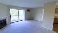 This single-floor space centers around a spacious, open living area finished with wall-to-wall carpeting in a neutral tone, creating a clean and understated aesthetic. The room is bathed in natural light, courtesy of a large sliding glass door that spans a significant portion of one wall. This door provides direct access to a private balcony enclosed by a white railing, which overlooks a verdant, tree-lined outdoor setting — a notable feature that brings a sense of openness and connection to nature into the interior.

The living area is well-proportioned, offering generous room for flexible furniture arrangements. A wall-mounted air conditioning unit is positioned above the sliding door, providing a dedicated climate control solution. The walls are finished in a warm beige tone that complements the neutral carpet and enhances the light, airy feel of the space.

An adjoining area is partially visible through a recessed opening, suggesting a connected room or entryway that adds to the overall flow of the layout. Neutral-toned baseboards frame the perimeter of the room, contributing to the clean, cohesive finish throughout. The overall ambiance is bright, quiet, and functional.