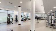 Located on Stadium Way in Madison, Alabama, this fitness center offers a comprehensive workout environment within a single-floor layout designed for both cardio and strength training. The space is characterized by a clean, modern aesthetic, with white walls, white structural columns accented by dark caps, and light-toned flooring that creates a bright, open atmosphere throughout.

Large windows line the perimeter walls, allowing ample natural light to filter in and providing views of the surrounding exterior. Recessed lighting and ceiling fans supplement the natural light, ensuring consistent illumination across all areas of the space.

The cardio zone features a row of treadmills equipped with individual digital screens, alongside elliptical trainers and stationary and upright bikes. A dedicated strength training area includes a rack system stocked with dumbbells in a full range of weights, a multi-functional cable machine, weight benches, and a power rack. Floor-to-ceiling mirrors along one wall visually expand the space while offering functional feedback during workouts.

An additional functional training area near the entrance showcases a storage rack with resistance bands, medicine balls, and other accessories, set against a decorative textured accent wall. Green-framed glass entry doors add a polished architectural detail.