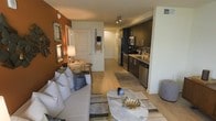 This single-floor residence showcases an open-concept layout that seamlessly connects the living area and kitchen into one cohesive, functional space. The living area is anchored by a plush white sofa adorned with decorative pillows and paired with a contemporary marble-top coffee table, creating an inviting and elegant focal point. A striking burnt-orange accent wall adds warmth and character, complemented by decorative mirrors that enhance the sense of depth in the room. A small desk and lamp tucked nearby offer a practical workspace without disrupting the overall flow.

Light wood-style flooring runs throughout, tying the spaces together while lending a clean, modern aesthetic. The kitchen features dark cabinetry contrasted by light countertops and a mosaic tile backsplash, giving it a sophisticated, contemporary feel. Stainless steel appliances, including a refrigerator, dishwasher, and microwave, provide a polished, functional finish. Track lighting above the kitchen ensures the workspace is well-illuminated. A hallway visible from the living area leads to additional spaces, contributing to the unit's overall sense of thoughtful layout and practical design.